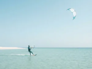 Beginner kitesurfer glijdt over kalm turkoois water met kite in heldere lucht, zandstrand op de achtergrond