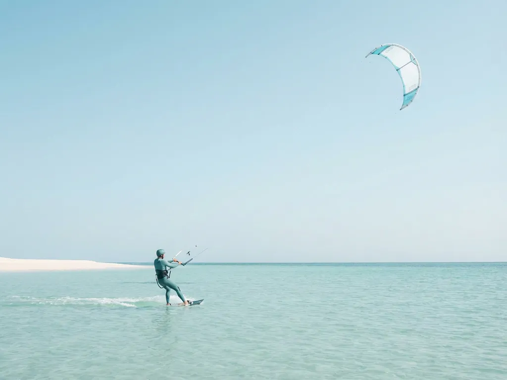 kitesurfen-beginner-turquoise-water-kalme-zee.jpg Beginner kitesurfer glijdt over kalm turkoois water met kite in heldere lucht, zandstrand op de achtergrond